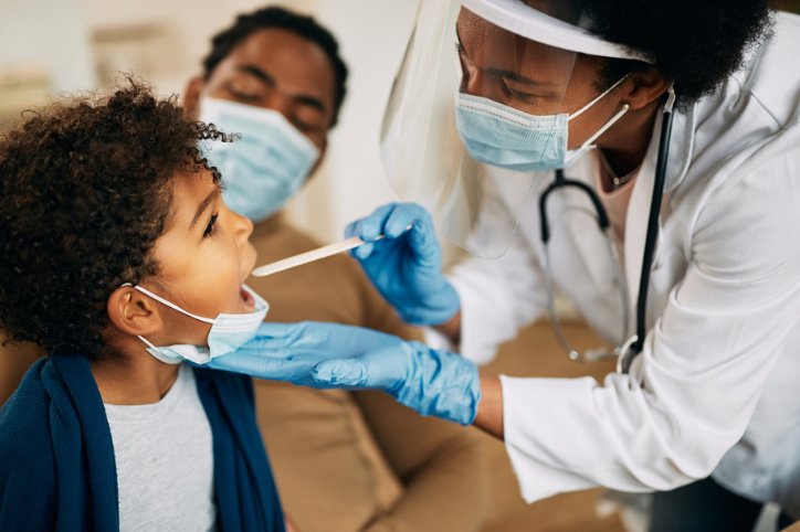A masked provider uses a tongue depressor to look into a happy child's throat