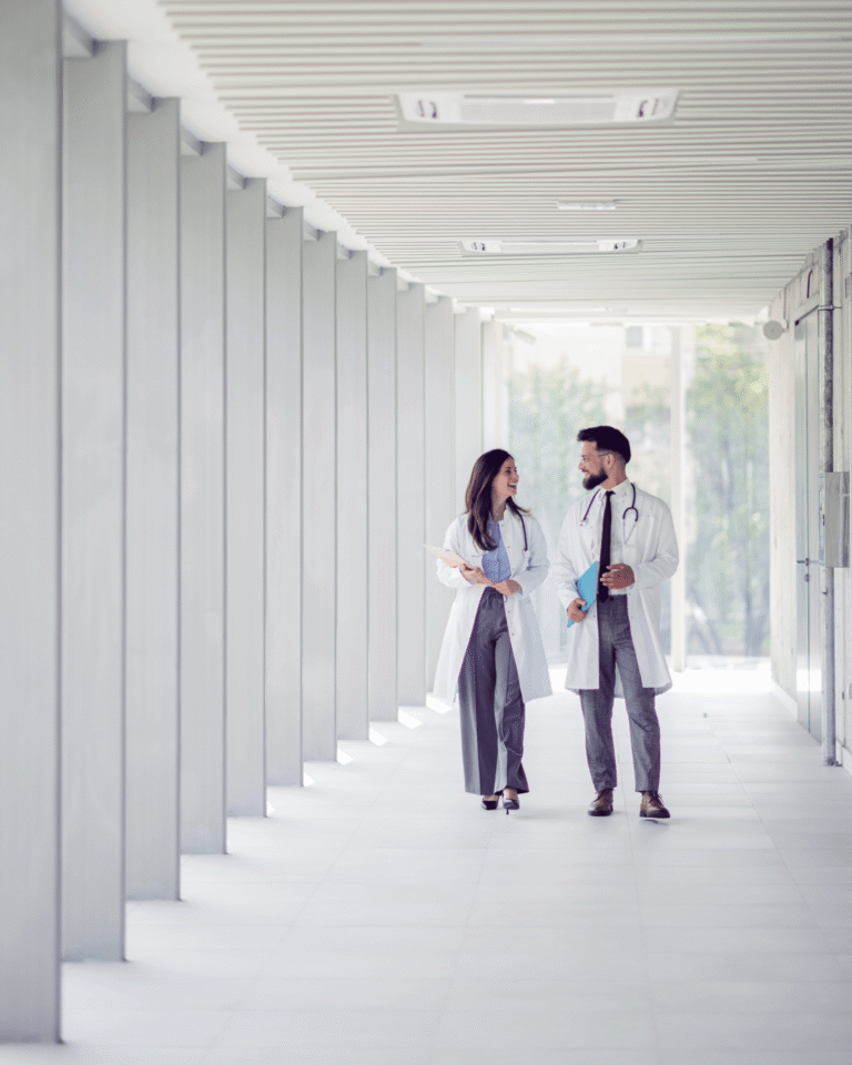 Two physicians, one man and one woman, walking together in a sunlit hospital hallway, smiling in conversation.